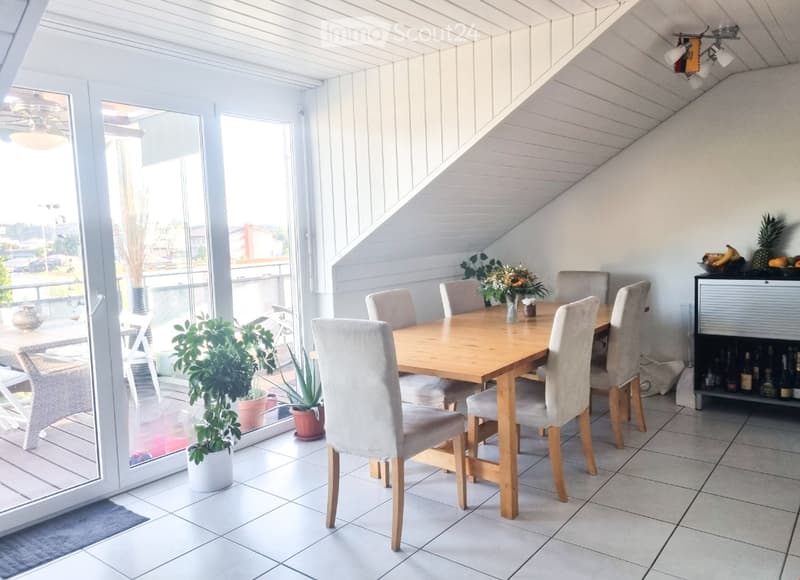 Bright dining area with a wooden table, six chairs, potted plants, and a bottle rack. Large windows and sliding doors open to a balcony.