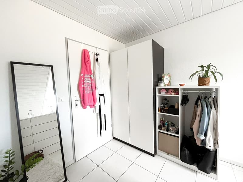 Modern, minimalist walk-in closet with white walls, tiled floor, large mirrored wardrobe, open shelving unit with clothes and accessories, and a potted plant.