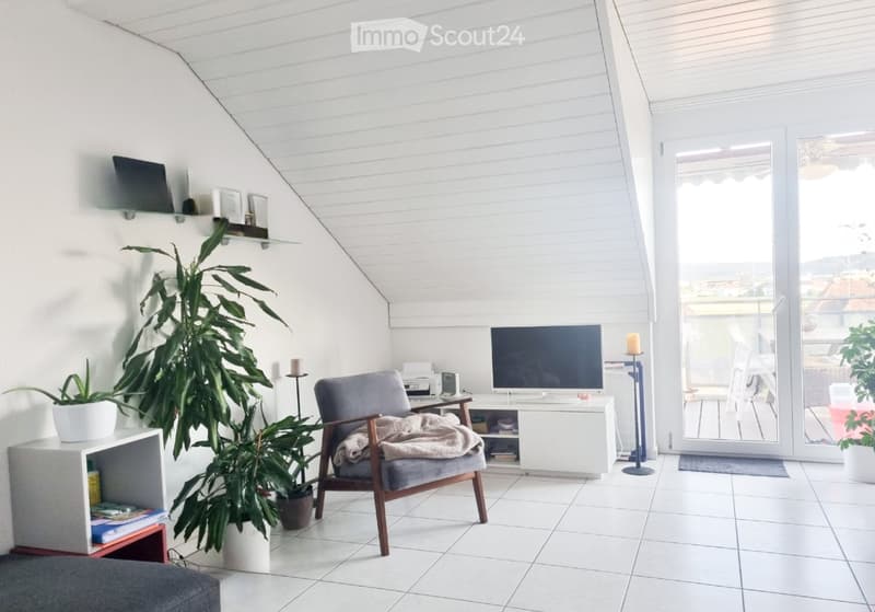Bright living room with white walls, tiled floor, large window, and a view. Features a grey armchair with a blanket, a TV on a white stand, a shelf with books and decorative items, and several potted plants.