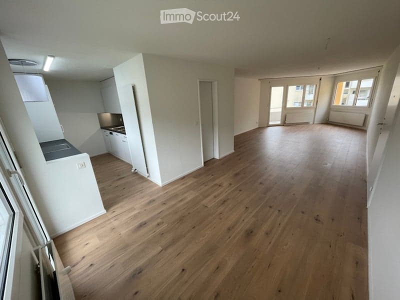 Empty room with wooden flooring, white walls, and a small kitchen area with cabinets and a sink.