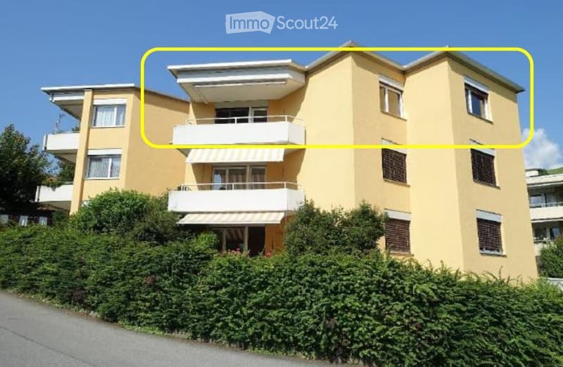 A modern, three-story residential building with beige walls, multiple balconies, and large windows. The building is surrounded by greenery and has a clean, contemporary design.