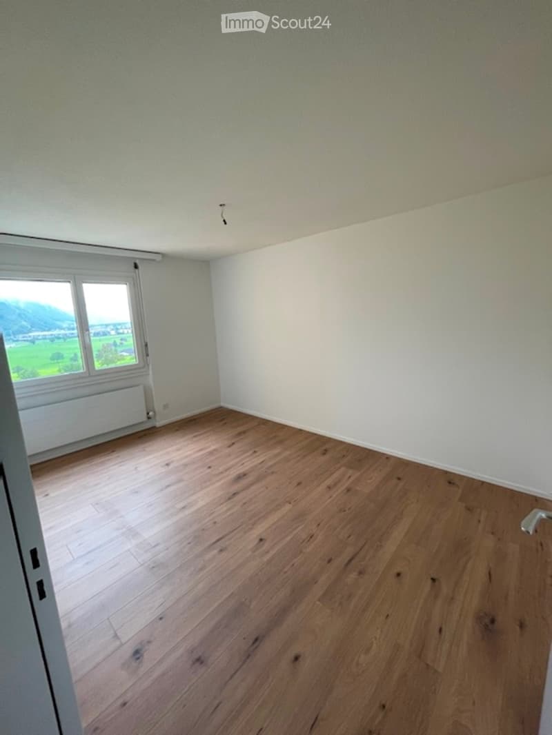 Completely empty room with wooden flooring and white walls, featuring a small window with a view of greenery outside.