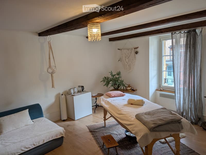 Cozy room with exposed wooden beams, a massage table, a sofa, a small table, a refrigerator, and a hanging light fixture.