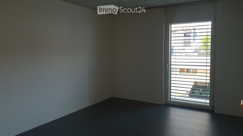 Empty room with bare walls and a grey floor, featuring a large window with blinds that offers a view of the outside.