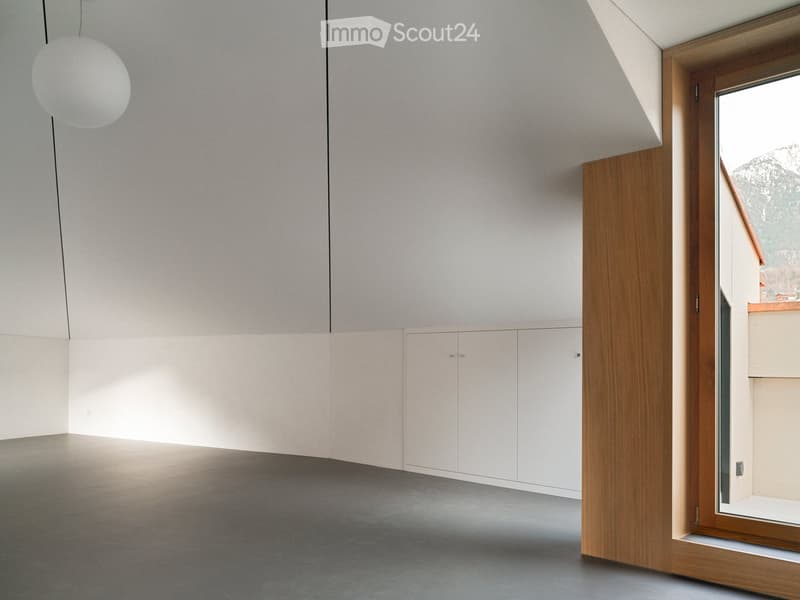 Empty room with grey walls, white built-in cabinets, and a large window with wooden frame. The floor is grey concrete.