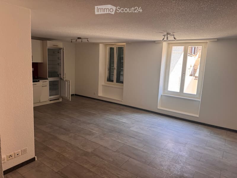 Empty room with tiled floor, white walls, and large windows with shutters. The room has built-in cabinets and a small kitchen area with a refrigerator and oven, but no furniture or personal items are present.