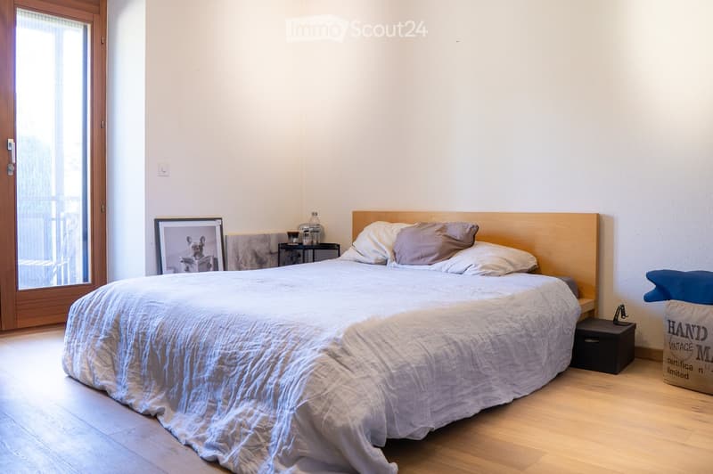 A modern bedroom with a large bed, nightstand, picture frame, and a bottle on the nightstand. The room features wooden flooring and a sliding glass door leading to a balcony.