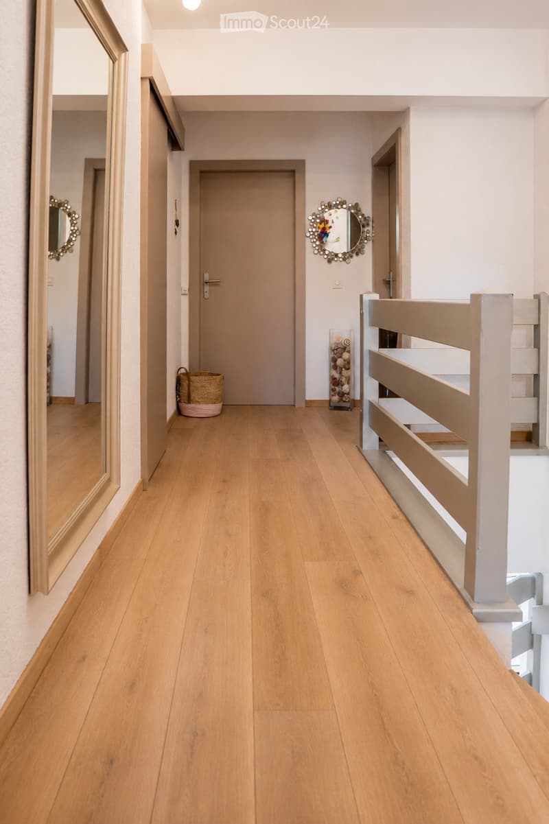 A clean, modern hallway with light wooden flooring, white walls, and a staircase with a white railing. Mirrors and decorative items are visible on the walls.