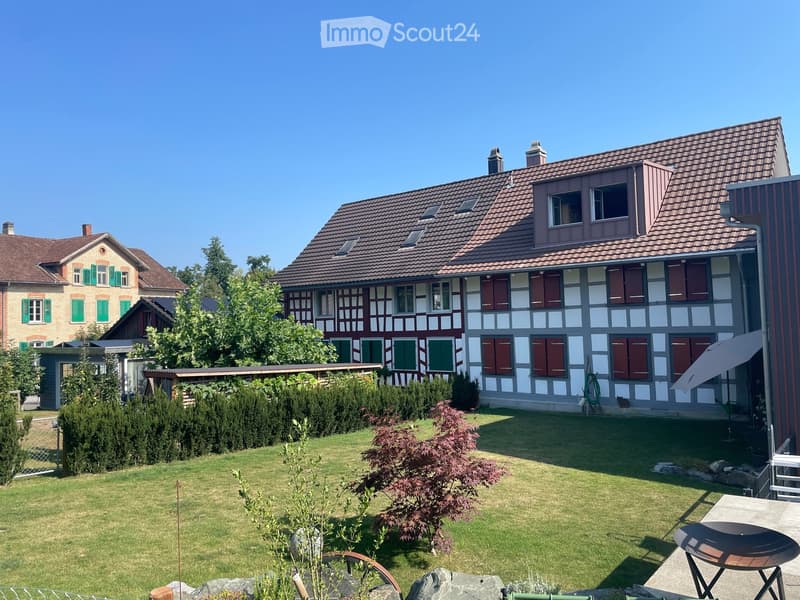 Well-maintained garden with a lawn, shrubs, and a small patio area featuring a table, chairs, and an umbrella. The house in the background has a traditional half-timbered design with red shutters and a tiled roof.