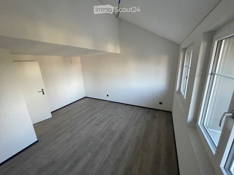 Empty room with light grey walls, wooden flooring, and two windows allowing natural light. The room features a white door on the left side and a small electrical outlet on the wall.