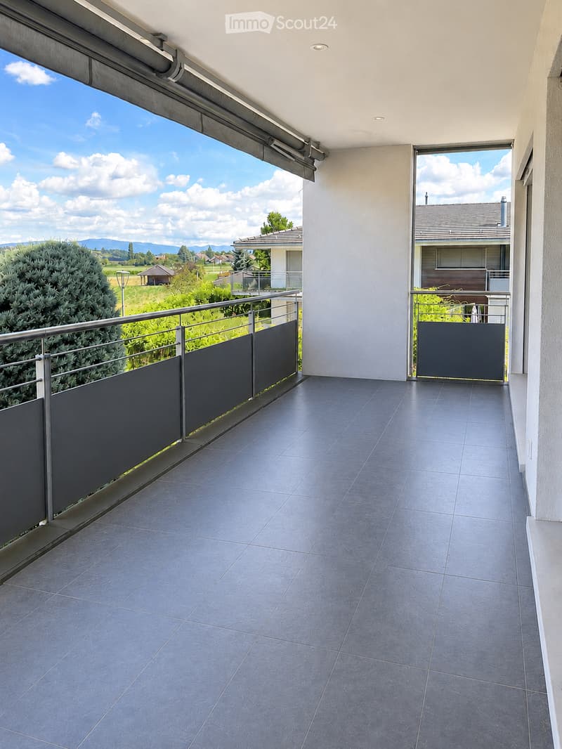 Balcony with grey tiled floor, metal railing, and retractable blinds, overlooking a green landscape with trees and distant mountains.