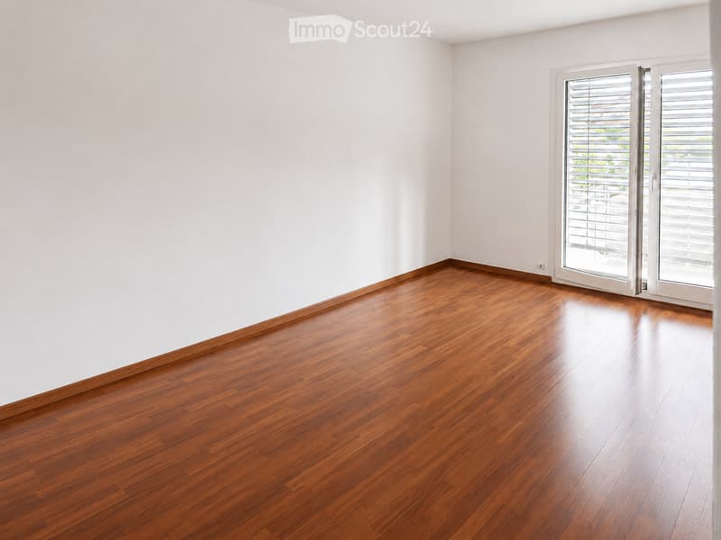 Empty room with light-colored walls and polished wooden flooring, featuring a large sliding glass door with blinds.