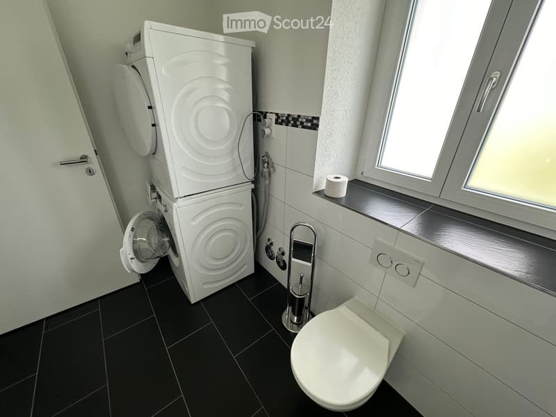 Modern bathroom with black tiled floor, white toilet, stacked washing machine, towel rack, and window with toilet paper on the sill.