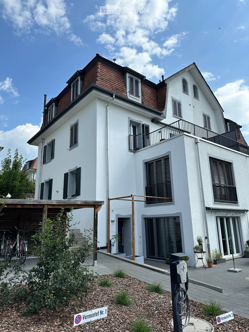 Modern two-story house with white exterior, red-tiled roof, multiple windows, balconies, and a covered garage area with bicycles.