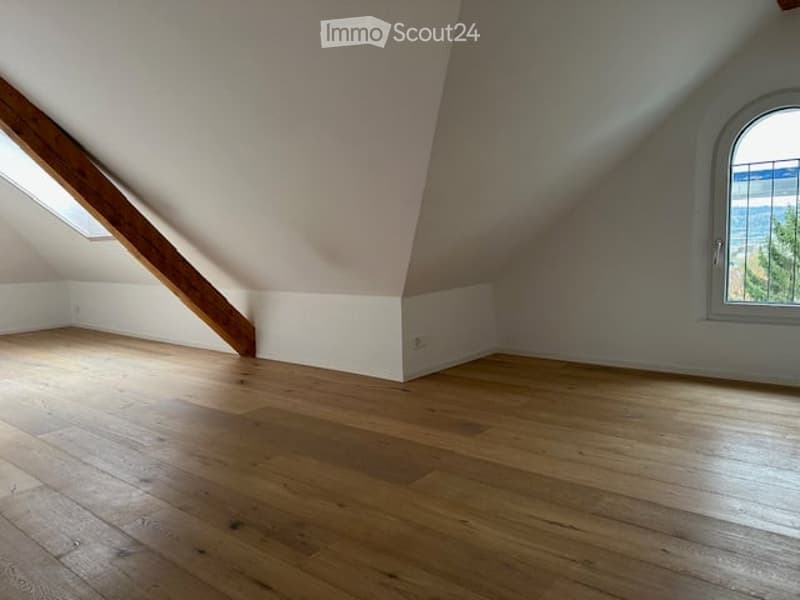 Empty attic room with wooden flooring, slanted ceilings, and a window offering a view.