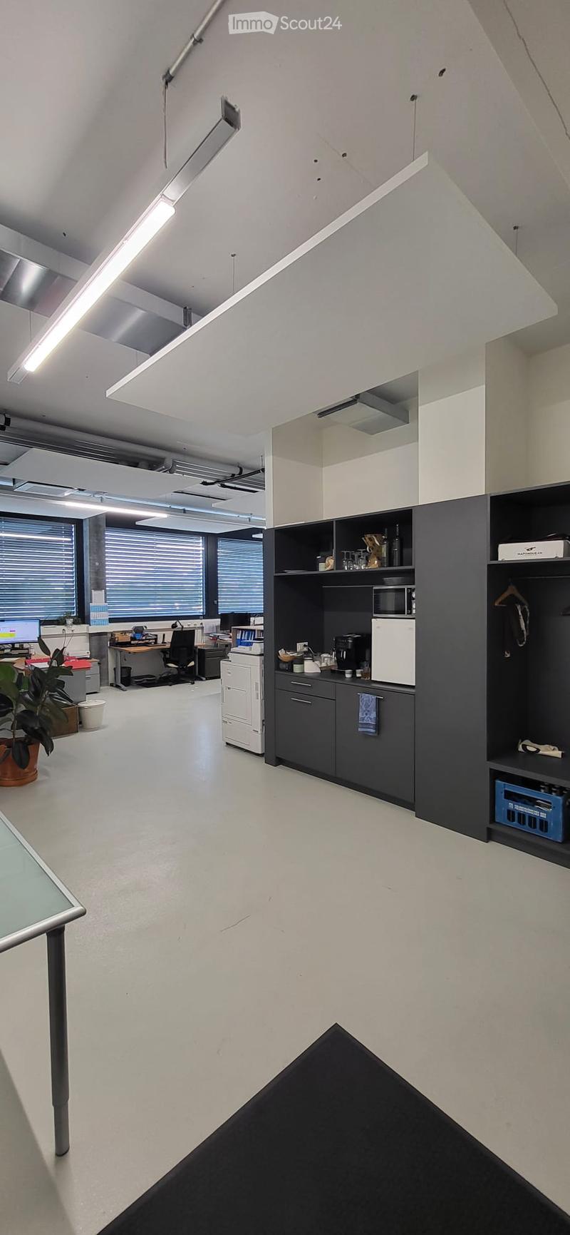 Modern office space with white walls, large windows, and a small kitchenette area. Includes desks, chairs, a printer, a microwave, and a coffee maker.