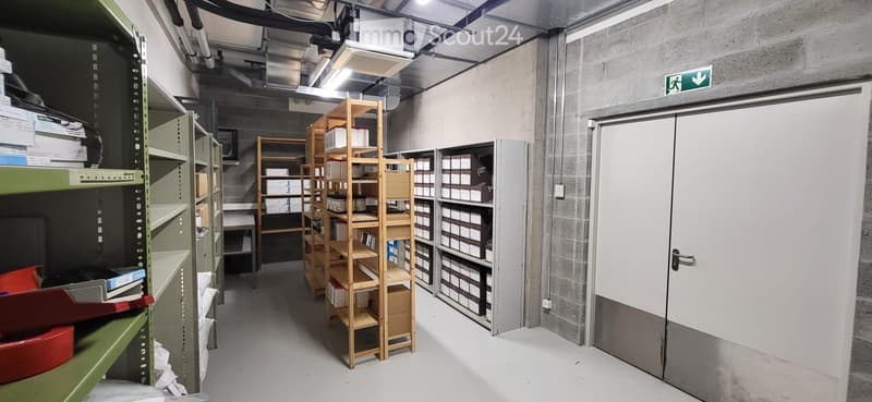 A storage corridor with metal shelving units, wooden shelves, and various boxes and items stored throughout. The space features concrete walls, a white floor, and an emergency exit sign above a door.