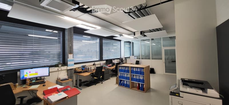 Modern office space with desks, chairs, computer monitors, filing cabinets, and a printer. Large windows with blinds provide natural light.