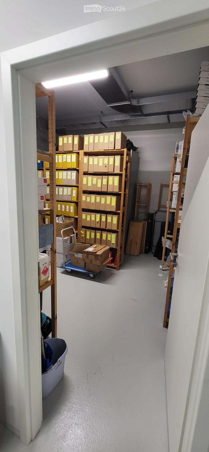Empty storage room with wooden shelving units, cardboard boxes, and a small cart. The room has white walls and a concrete floor.