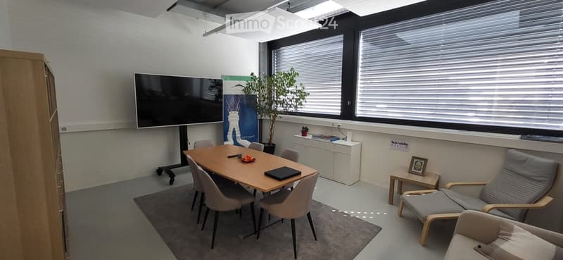 Modern office or meeting room with a large table, chairs, a sofa, a screen, a cabinet, and a plant. The room has large windows with blinds and a neutral color scheme.