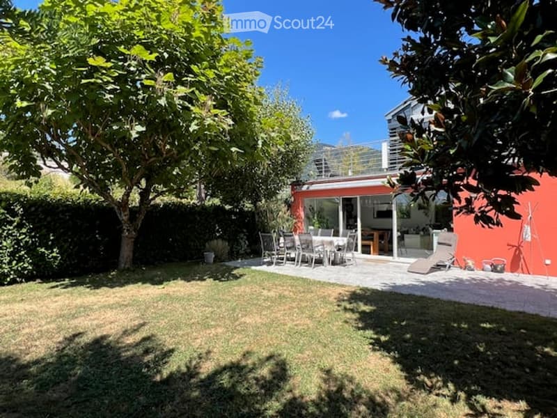 A well-maintained garden with a lawn, trees, and a patio area featuring a dining set and lounge chair. The patio is adjacent to a modern house with large glass doors.