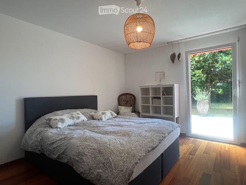 Bright bedroom with wooden flooring, large window, and sliding glass door leading to a balcony. Features a bed with pillows and a blanket, a nightstand, a chair, and a bookshelf.