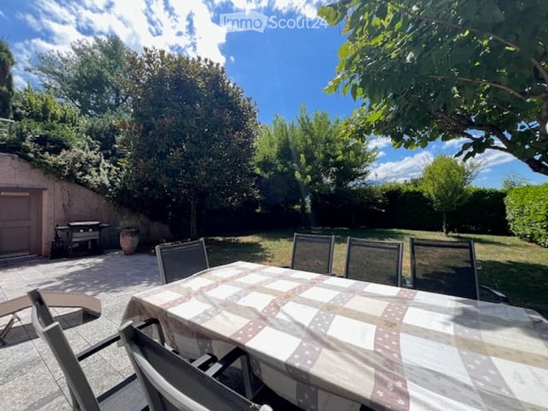 Outdoor patio area with a dining table, chairs, and lounge chairs, surrounded by trees and greenery.