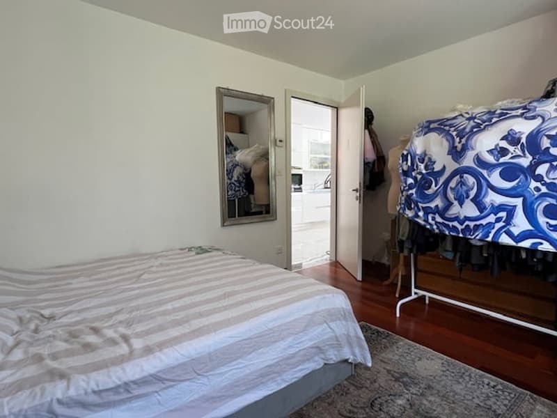 Cozy bedroom with a double bed, striped duvet, wooden floor, large mirror, and open closet with clothing items.