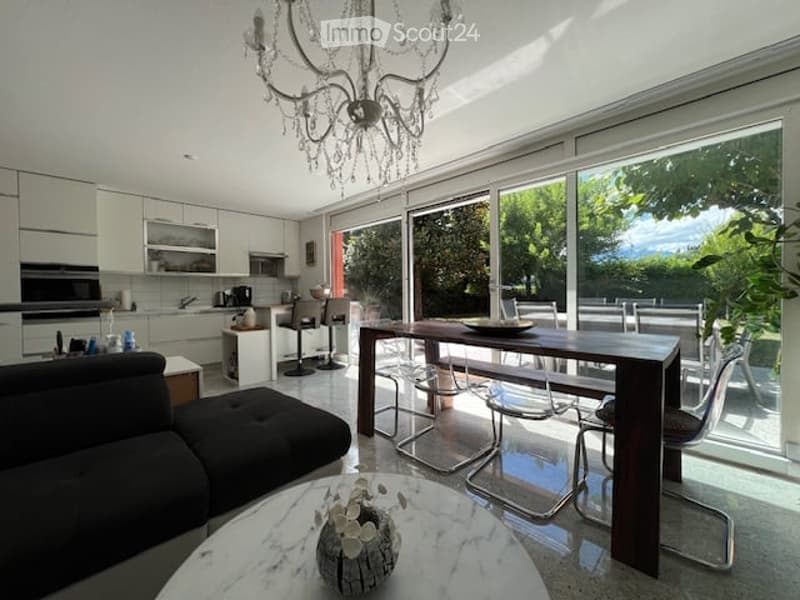 Bright, modern open-plan living and dining area with large glass windows, a black sofa, a round marble coffee table with a decorative vase, a dark wooden dining table with modern chairs, and a sleek white kitchen with cabinets, a stove, and a sink.