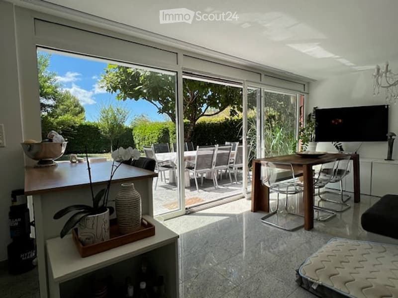 Bright, modern living space with large sliding glass doors opening to a patio, dining area with a wooden table and chairs, a counter with decorative items, and a television mounted on the wall.