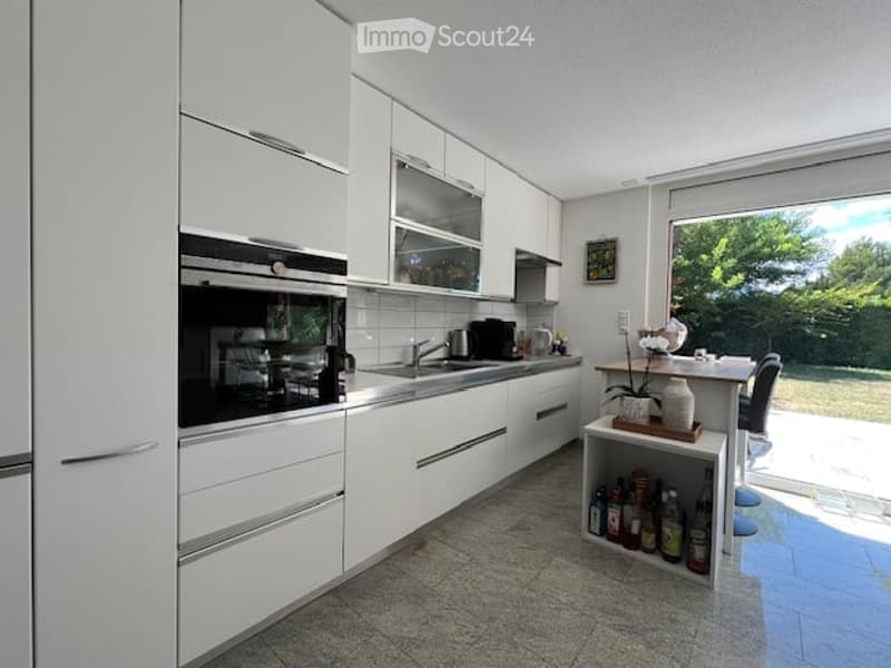 Modern kitchen with white cabinets, stainless steel appliances, and a large window overlooking a garden. Features include an oven, sink, coffee maker, and a small dining area with a table and chairs.