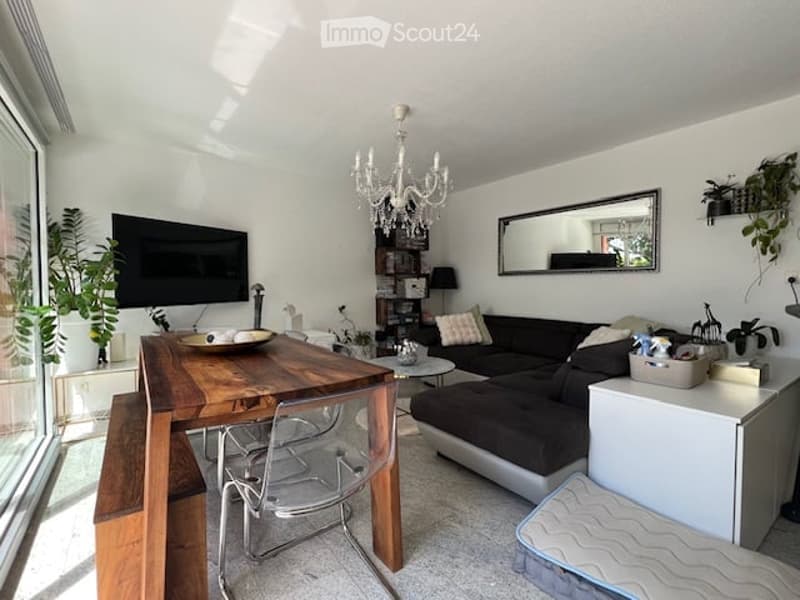 Bright living room with white walls, a dark grey sectional sofa, a wooden dining table with chairs, a chandelier, a TV, and several plants.