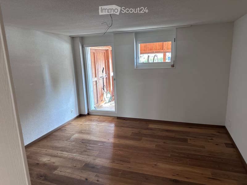 Empty room with wooden flooring, white walls, a window with blinds, and an open door leading outside.