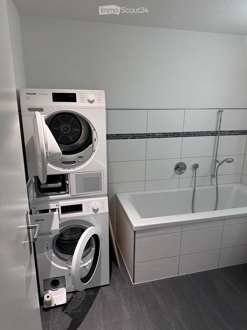 Modern bathroom with a bathtub, washing machine, and dishwasher. The washing machine and dishwasher are stacked vertically, with their doors open, revealing the interior. The bathtub is positioned next to the appliances, and the walls are tiled in light g