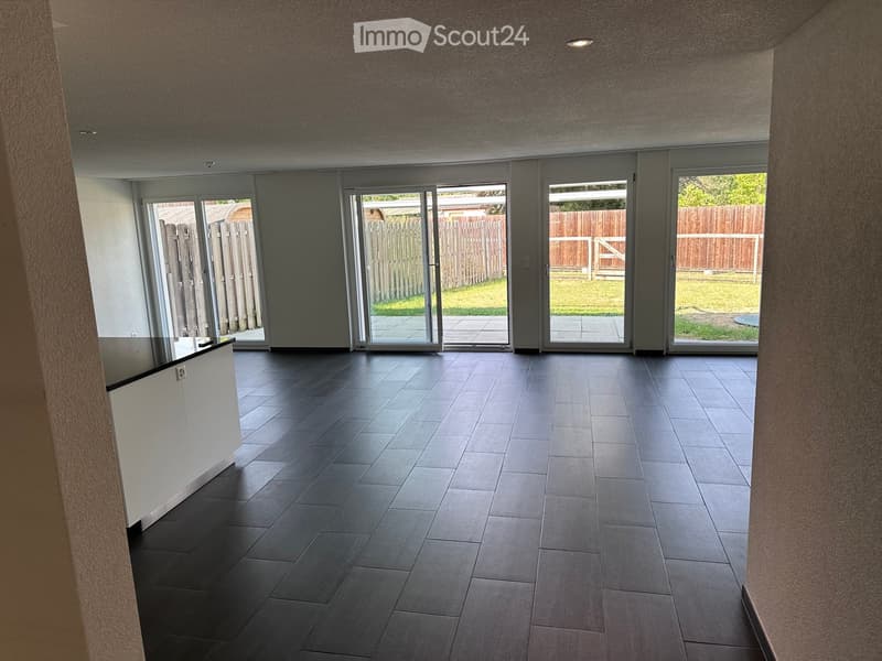An empty room with dark tiled flooring, white walls, and large sliding glass doors leading to an outdoor area with a wooden fence and grass.