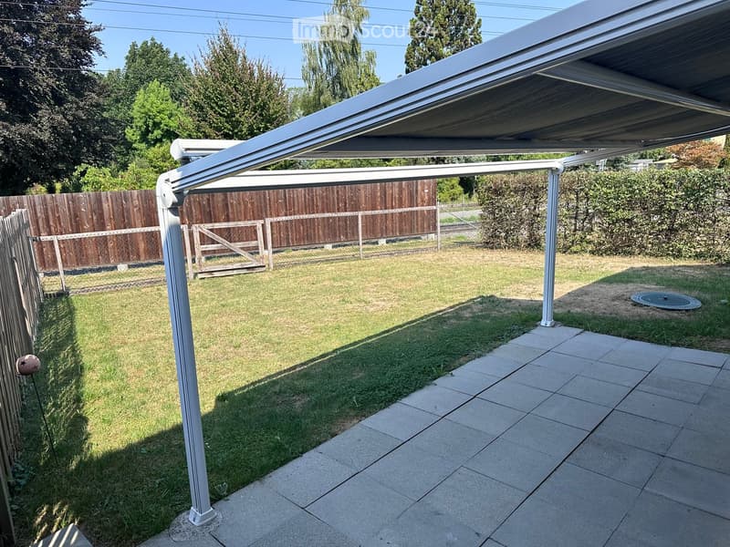A covered patio area with a retractable roof, surrounded by a wooden fence, grass, and trees.