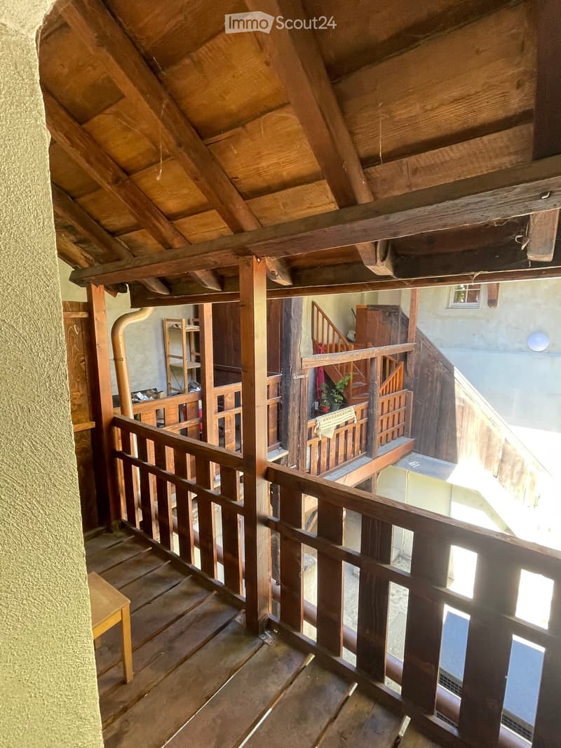 Wooden staircase with wooden railings and steps, leading to an upper level with visible wooden beams and exposed wooden ceiling structure. The walls are made of stone and wood, and there is a small wooden bench on the left side of the staircase.