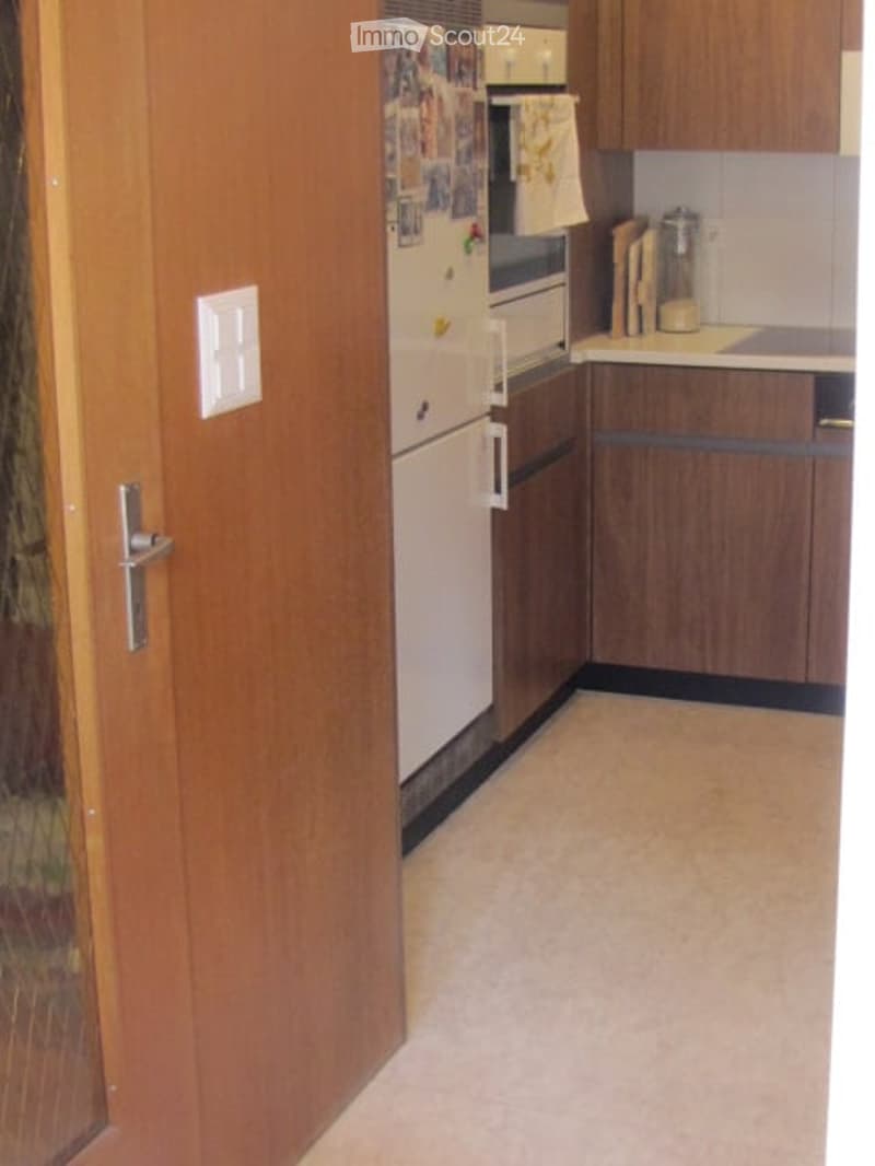 A small kitchen with wooden cabinets, a white refrigerator, and a countertop with a few items including a cutting board and a jar. The floor is light-colored tile.