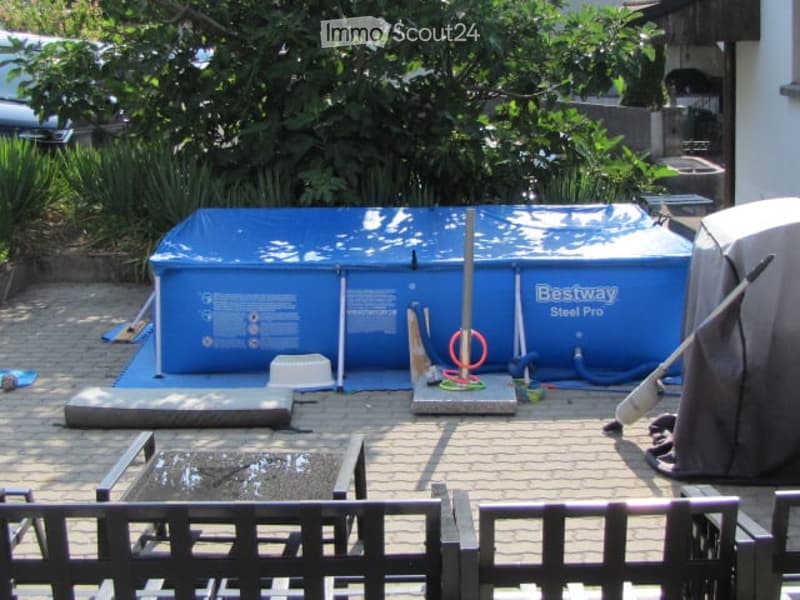 Backyard with a blue inflatable swimming pool, surrounded by a wooden deck, various pool accessories, and greenery.
