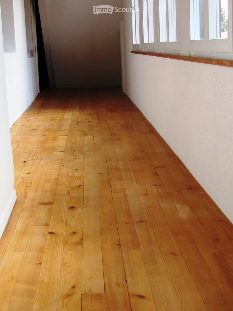 Empty hallway with polished wooden flooring and white walls, featuring a window with white frames and a small wooden shelf above it.