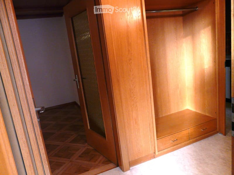 Empty room with wooden floor, open wooden closet with shelves and drawers, and a glass-paneled door leading to another area.