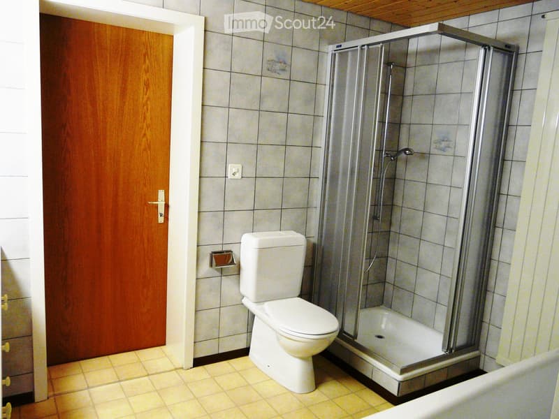 Bathroom with a toilet, a glass-enclosed shower stall, tiled walls and floor, and a wooden door.