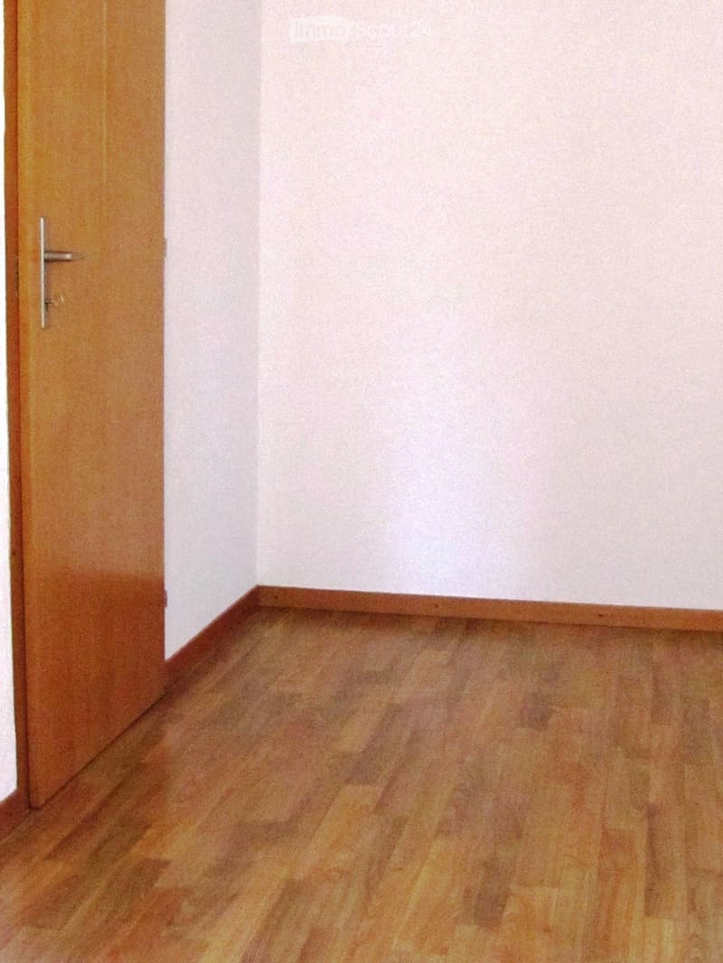 Empty room with wooden flooring and white walls, featuring a wooden door with a silver handle on the left side.