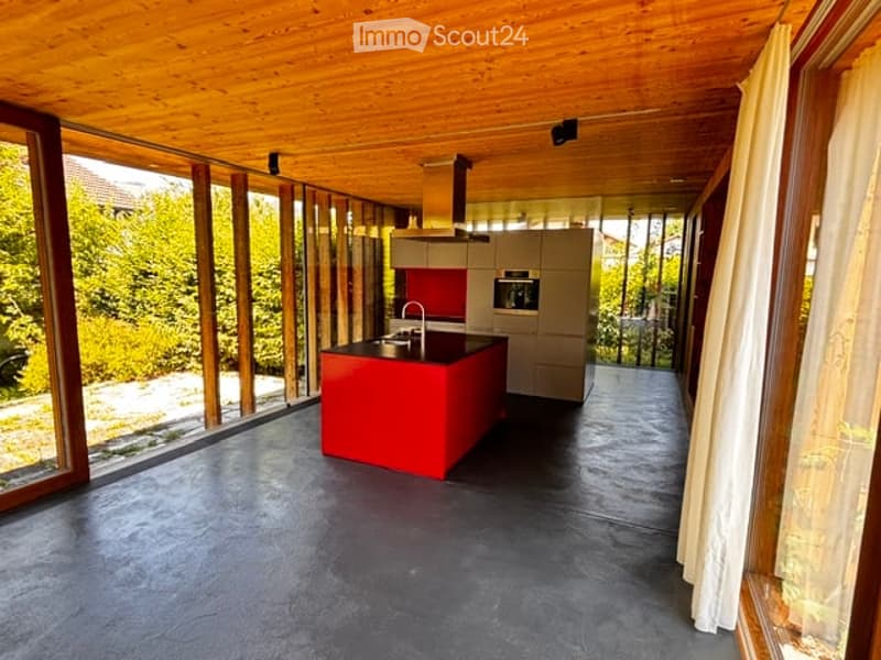 Modern kitchen with a red island, stainless steel sink, built-in oven, and large windows offering a view of the greenery outside.