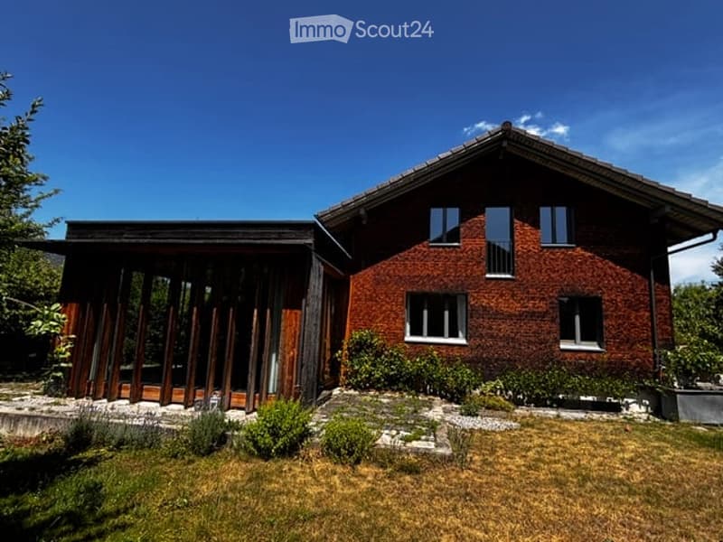 Two-story house with red brick facade and large windows, featuring a wooden deck and landscaped garden.