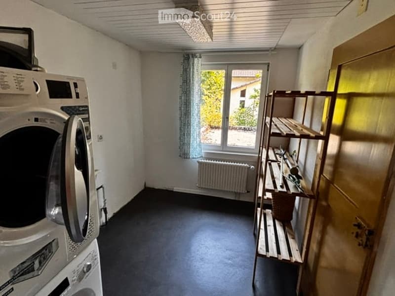Small utility room with a washing machine, shelf, window with curtain, and radiator.