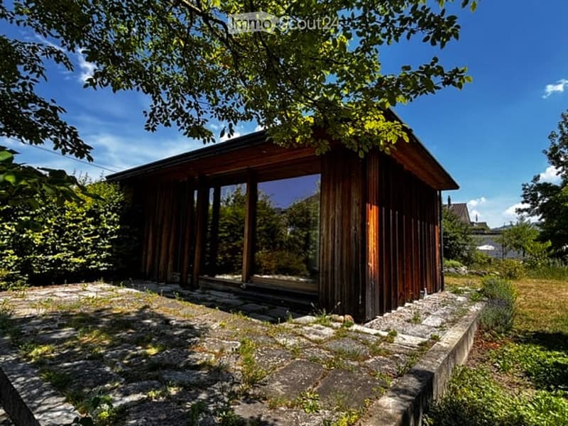 A small wooden cabin with large glass windows, surrounded by greenery and a stone pathway.