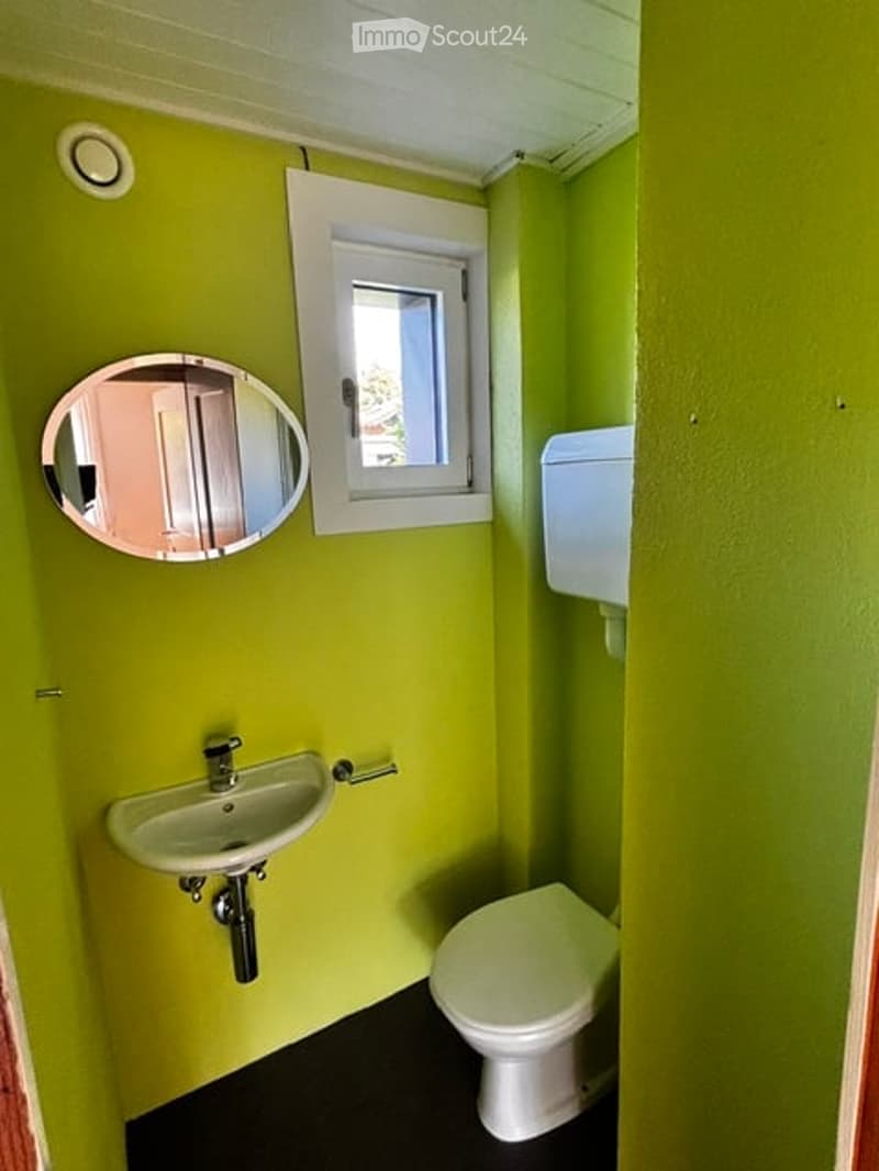 Bright green bathroom with a small window, white sink, toilet, and round mirror.