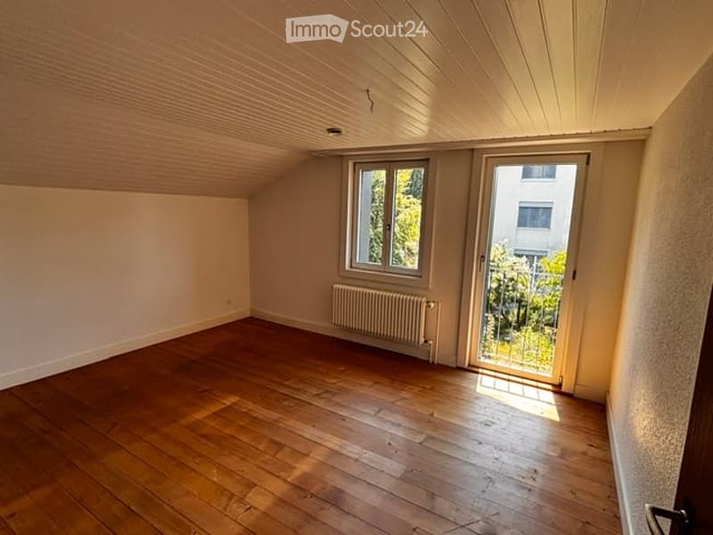 Empty attic room with wooden flooring, white walls, and large windows offering a view of greenery outside.