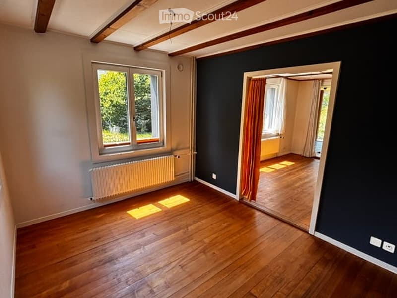 Empty room with wooden flooring, large window with radiator below, dark blue accent wall, and open doorway leading to another room.